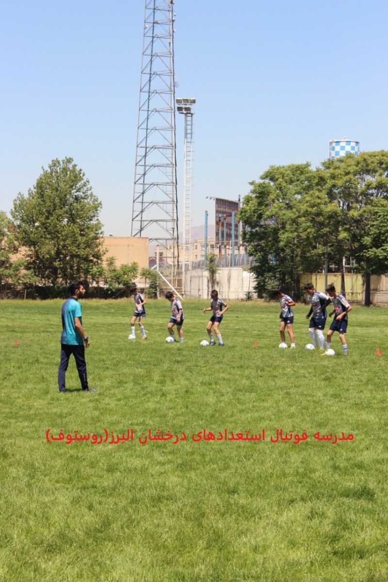 تصویری از زمین چمن مدرسه فوتبال استعدادهای درخشان البرز (روستوف)، بهترین مدرسه فوتبال در کرج، با بازیکنان جوان در حال تمرین زیر نظر مربی، با لباس‌های ورزشی، در کنار برج و درختان